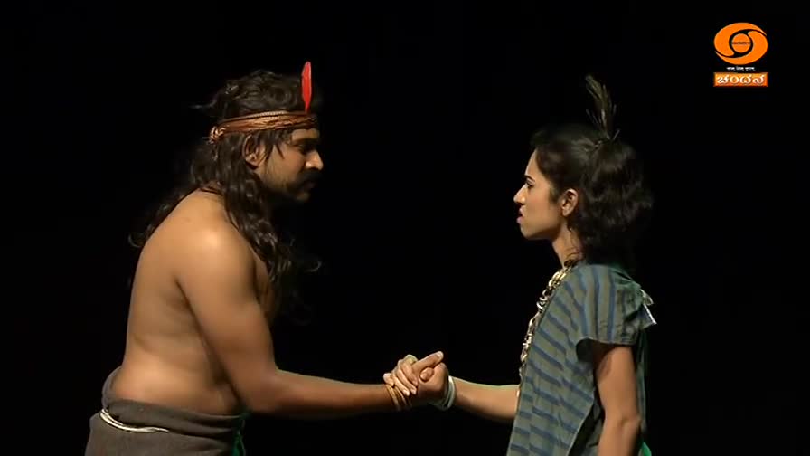 A man with long hair and a headband clasps hands with a woman wearing a striped top, their faces close. The DD Chandana logo is visible in the corner, suggesting a performance.

