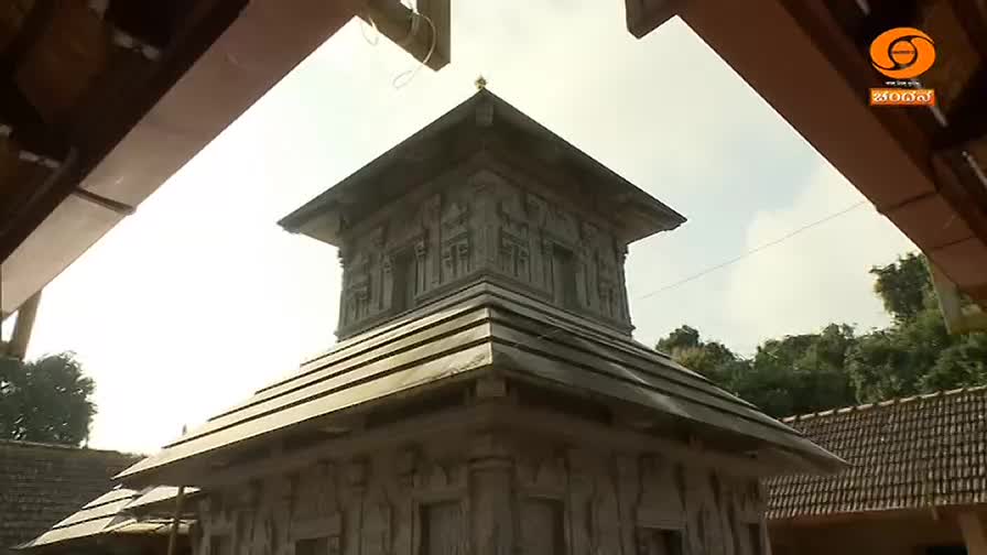 A stone temple, its roof layered like steps, rises against the bright sky. Through the frame of a wooden structure, the DD Chandana logo sits in the corner.
