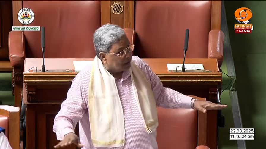 A man in a pink striped shirt gestures with both hands, speaking into a microphone. He's wearing a white scarf and glasses, and the DD Chandana logo is visible on the screen.

