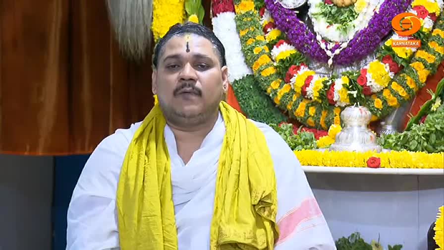 The man in the white robe and yellow shawl is speaking, his gaze directed towards the camera. Behind him, a decorated altar with flowers and religious items is visible.
