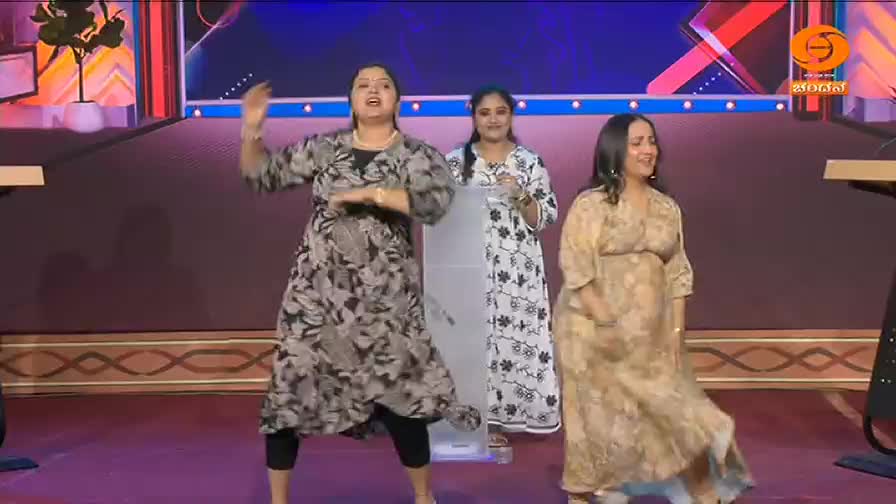 Three women on a stage are dancing, one in a floral print dress is leading the movement. The DD Chandana logo is visible in the corner.

