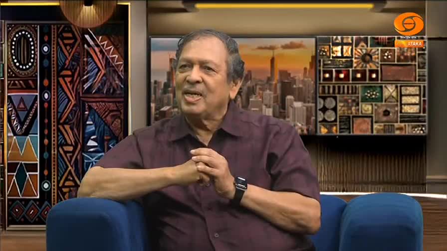A man in a maroon shirt sits in a blue armchair, speaking with his hands clasped. Behind him, the set of what appears to be a DD Chandana show features a cityscape backdrop and decorative panels.
