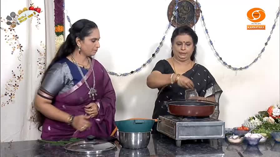 On a DD Chandana set, one woman in a black sari pours something from a small glass into a pan on a burner. The other woman in a purple sari watches, her hands resting on the countertop.
