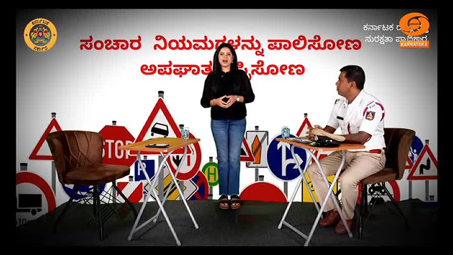 A woman in jeans and a black top stands center stage, gesturing as she speaks. A traffic officer sits to her right, listening attentively at a small table, with road signs as a backdrop.
