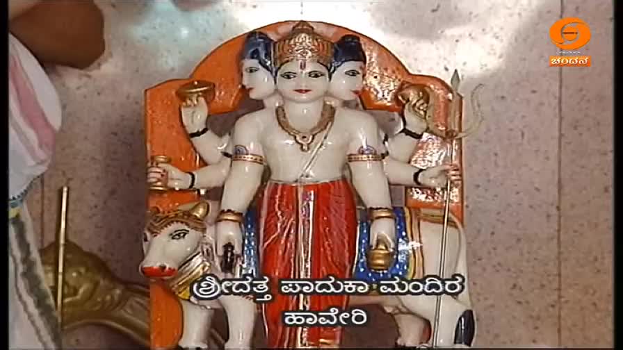 A multi-faced statue of a deity stands centered, its white marble body contrasting with the red and gold of its clothing. The DD Chandana logo is visible in the corner, suggesting this scene is part of a broadcast from India.
