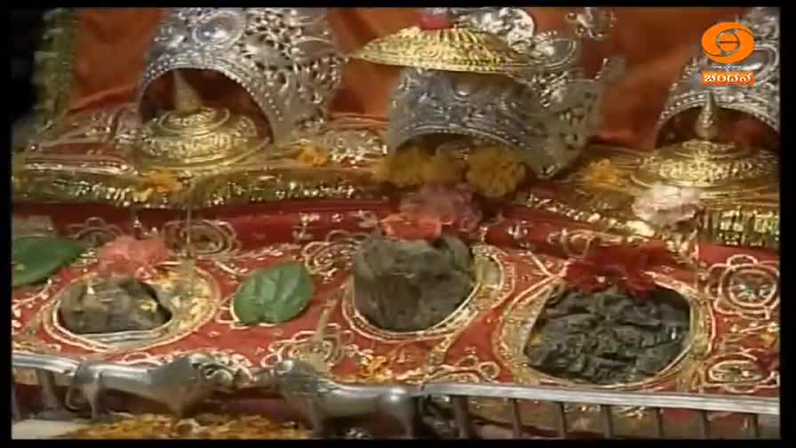 Ornate silver and gold adornments sit atop a red cloth, framing dark, rounded stones. Scattered flowers and green leaves add a touch of color to the scene.
