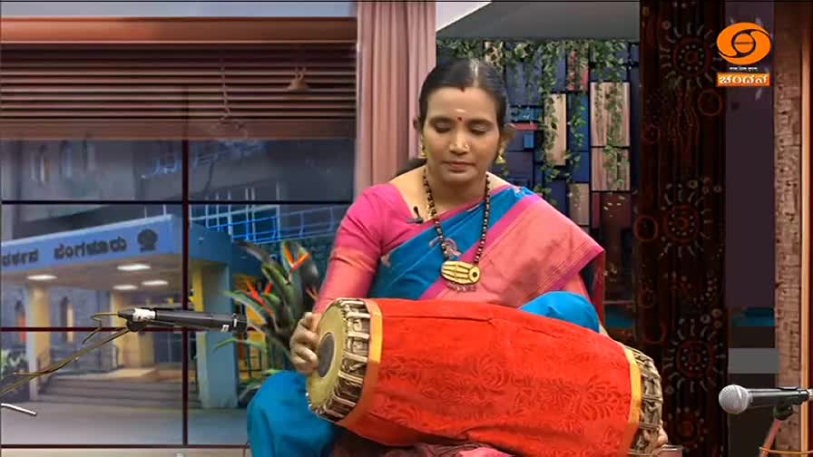 A woman in a vibrant saree sits cross-legged, cradling a large, red mridangam. Her hands move rhythmically, striking the drum as she performs on DD Chandana, a television channel from India.
