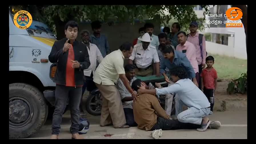 A man lies on the ground, being attended to by several people, while a news anchor from DD Chandana gestures towards the scene. A crowd gathers around, including a police officer and a small boy in a red shirt.
