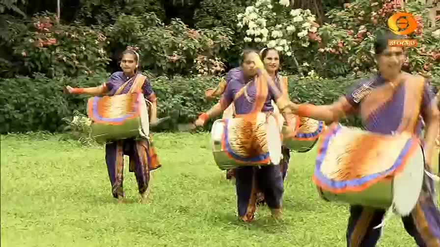 Dancers in vibrant saris move across the grass, each carrying a large, decorated drum. Their arms swing rhythmically as they perform, the drums bouncing gently with their steps.
