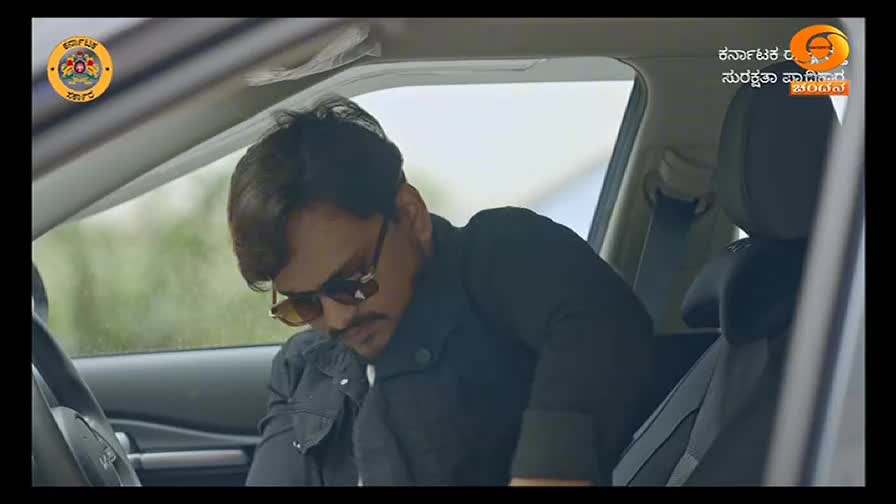 A man in sunglasses is inside a car, leaning forward. He seems to be looking at something in his lap, perhaps a phone or papers.
