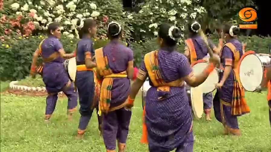 Dancers in purple and orange saris move in a circle, some holding drums. The rhythmic beat of the drums and the dancers' steps fill the air.
