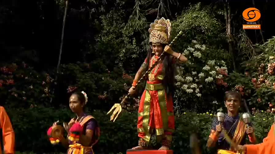 A performer, costumed as a deity, stands on a small platform, holding a trident high. Two women play small cymbals and drums, while others hold instruments, all part of a traditional performance.
