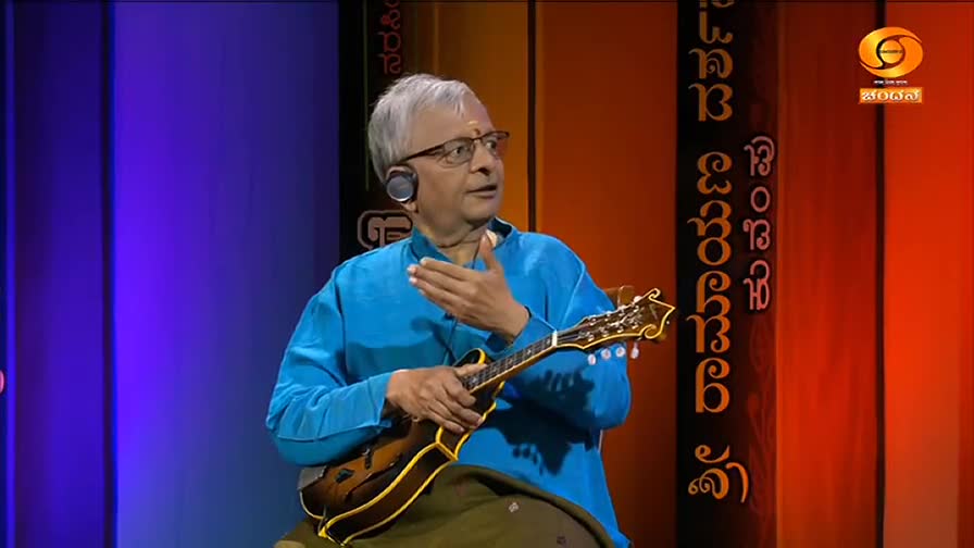 A man in a blue kurta gestures with his right hand while holding a mandolin. He's wearing glasses and a headset, likely on the DD Chandana set in India.
A man in a blue kurta gestures with his right hand while holding a mandolin. He's wearing glasses and a headset, likely on the DD Chandana set in India.
