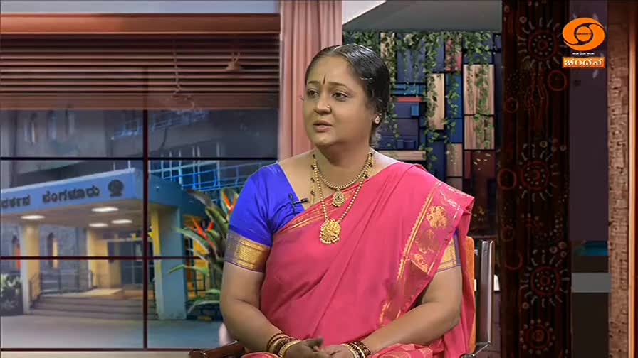 A woman in a pink sari and gold jewelry sits and speaks, framed by a window showing a building. The DD Chandana logo is visible in the corner.
A woman in a pink sari and gold jewelry sits and speaks, framed by a window showing a building. The DD Chandana logo is visible in the corner.