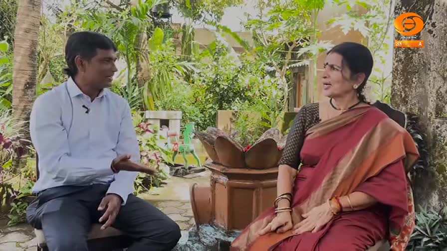 A man in a light blue shirt gestures as he speaks, while a woman in a red sari listens intently. The scene, set outdoors, is likely a DD Chandana interview.
A man in a light blue shirt gestures as he speaks, while a woman in a red sari listens intently. The scene, set outdoors, is likely a DD Chandana interview.