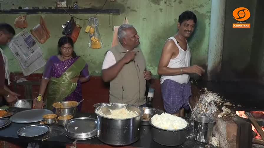 A man in a white tank top stirs something over a wood fire, while others stand around a table laden with bowls and plates. The DD Chandana logo is visible in the corner.
A man in a white tank top stirs something over a wood fire, while others stand around a table laden with bowls and plates. The DD Chandana logo is visible in the corner.