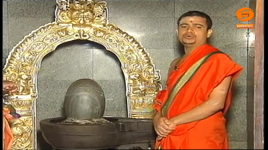A man in orange robes stands before a shrine, speaking directly to the camera. Behind him, a stone lingam sits in a basin, framed by an ornate golden arch.
A man in orange robes stands before a shrine, speaking directly to the camera. Behind him, a stone lingam sits in a basin, framed by an ornate golden arch.