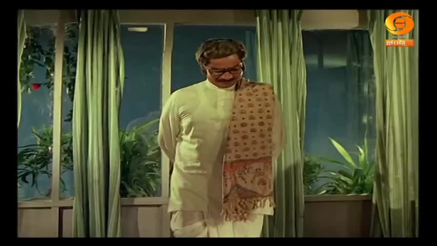 A man in a kurta and shawl stands in a room, his gaze fixed downwards. Behind him, the window reveals a glimpse of greenery and the logo of DD Chandana.
A man in a kurta and shawl stands in a room, his gaze fixed downwards. Behind him, the window reveals a glimpse of greenery and the logo of DD Chandana.