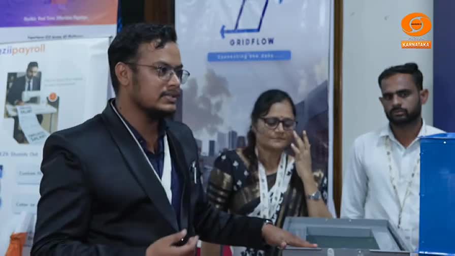 A man in a blazer gestures toward a device with a clear top, while two others look on. The backdrop includes a sign for "Gridflow" and a logo for Karnataka.
A man in a blazer gestures toward a device with a clear top, while two others look on. The backdrop includes a sign for "Gridflow" and a logo for Karnataka.
