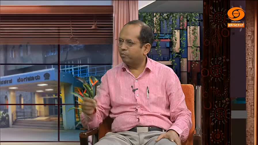 A man in a pink shirt gestures with a small bouquet of flowers as he speaks. Behind him, a window reveals a building with writing in Kannada, likely part of a DD Chandana broadcast from India.
A man in a pink shirt gestures with a small bouquet of flowers as he speaks. Behind him, a window reveals a building with writing in Kannada, likely part of a DD Chandana broadcast from India.
