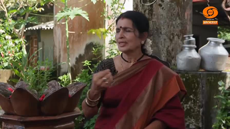 A woman in a red and gold sari gestures as she speaks, framed by lush greenery and a stone structure. The DD Chandana logo is visible in the corner, suggesting a broadcast from India.
A woman in a red and gold sari gestures as she speaks, framed by lush greenery and a stone structure. The DD Chandana logo is visible in the corner, suggesting a broadcast from India.