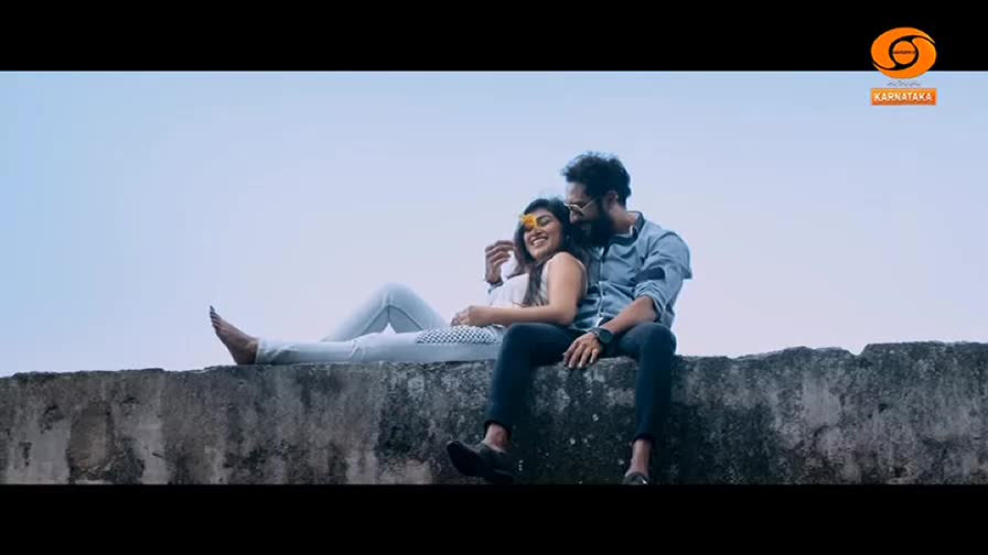 A couple sits on a concrete wall, the woman reclining with a flower in her hair. The man, with a beard and sunglasses, has his arm around her, and they both appear to be smiling.
A couple sits on a concrete wall, the woman reclining with a flower in her hair. The man, with a beard and sunglasses, has his arm around her, and they both appear to be smiling.