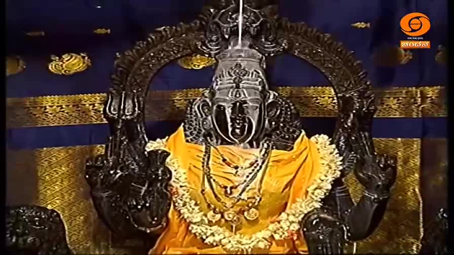 A dark, ornate statue of a deity is centered, draped in a yellow cloth and garlands. The statue is framed by an elaborate arch against a backdrop of blue and gold fabric.
A dark, ornate statue of a deity is centered, draped in a yellow cloth and garlands. The statue is framed by an elaborate arch against a backdrop of blue and gold fabric.