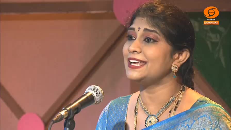 A woman in a blue sari speaks into a microphone, her face lit with a warm smile. Behind her, a logo for DD Chandana, a Kannada-language television channel in India, is visible.
A woman in a blue sari speaks into a microphone, her face lit with a warm smile. Behind her, a logo for DD Chandana, a Kannada-language television channel in India, is visible.