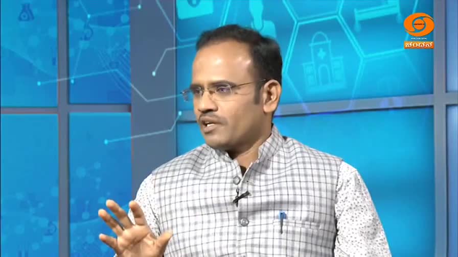 The man, wearing glasses and a checkered vest, gestures with his right hand as he speaks. A DD Chandana logo is visible in the corner, and the backdrop is a light blue with graphic elements.
The man, wearing glasses and a checkered vest, gestures with his right hand as he speaks. A DD Chandana logo is visible in the corner, and the backdrop is a light blue with graphic elements.