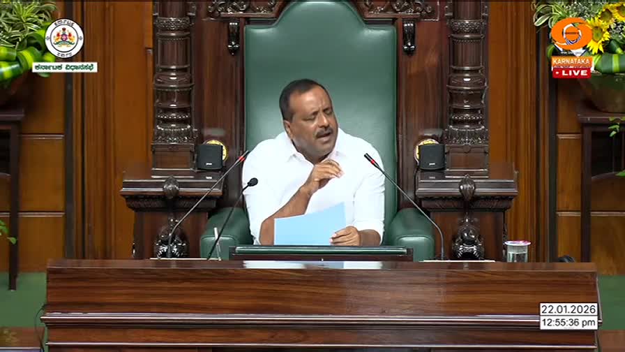 A man in a white shirt speaks from a large, ornate chair, holding a blue document. The Karnataka Legislative Assembly is in session, broadcast live. A man in a white shirt speaks from a large, ornate chair, holding a blue document. The Karnataka Legislative Assembly is in session, broadcast live.
