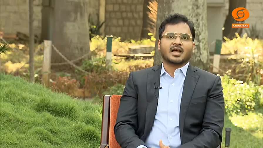 A man in a dark suit and light blue shirt speaks directly to the camera. Behind him, a lush green lawn slopes upwards, dotted with trees and a rustic fence. A small logo for DD Chandana Karnataka is visible in the upper right corner.