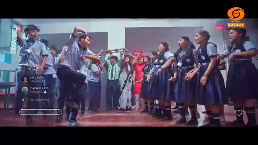 A group of students in uniform are dancing and singing on a stage, with a DD Chandana logo visible. A teacher in a sari stands behind them, smiling.