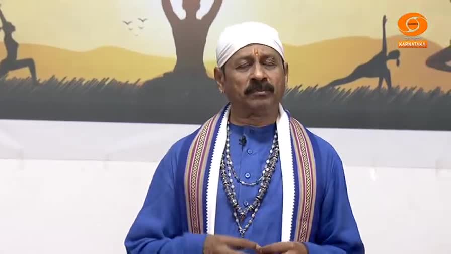 A man with a white turban and blue kurta stands with his eyes closed, a serene expression on his face. Behind him, a mural depicts figures in yoga poses against a sunset sky, with a DD Chandana logo visible.