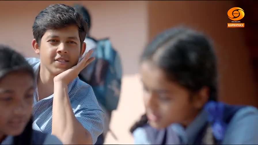 A young man in a blue shirt leans forward, his hand resting on his cheek, looking towards the camera. Behind him, other students are visible, their faces blurred. The DD Chandana logo appears in the corner.