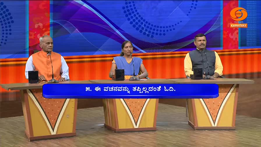 Three people sit behind a desk on a television set. A blue banner with Kannada text stretches across the front of the desk.