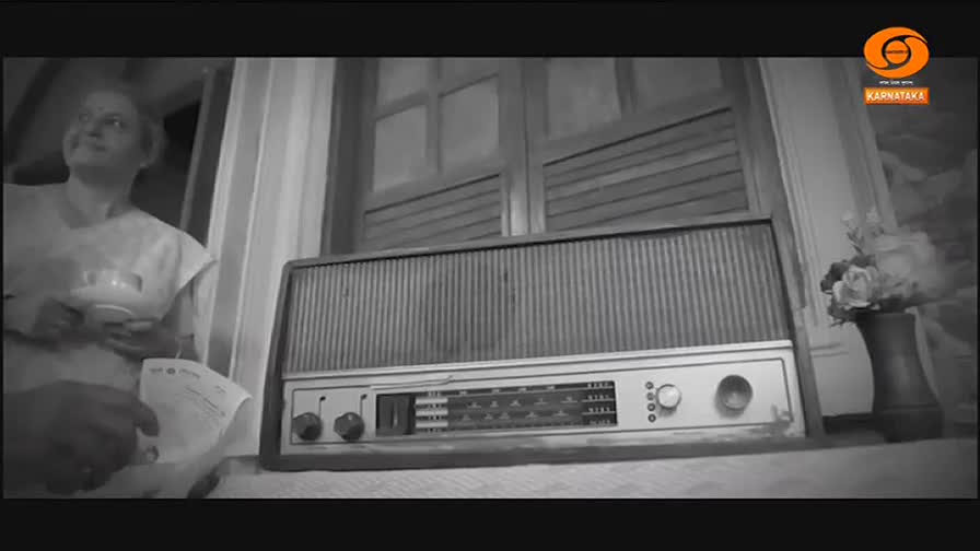 A woman in a light sari holds a cup and saucer, looking towards the window. Beside her, a vintage radio sits on a surface, its tuning dial illuminated.