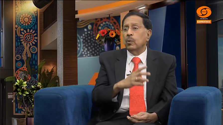 A man in a suit and bright red tie speaks animatedly, his hands gesturing. He's seated on a blue couch, likely on a DD Chandana program, with a colorful backdrop behind him.