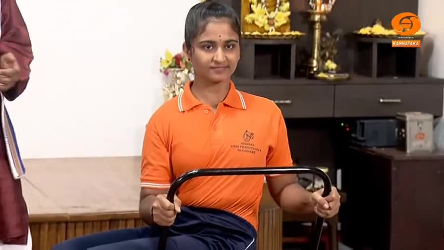 A young woman in an orange polo shirt grips a metal bar, her knuckles white. She's seated, her posture straight, as if preparing for an exercise demonstration on DD Chandana.