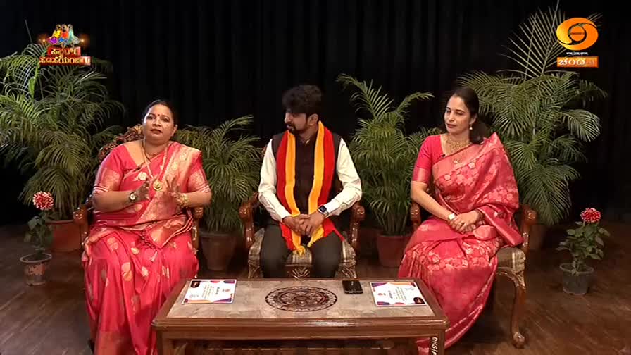 A woman in a pink sari gestures with her hands as she speaks. Beside her, a man wearing a white shirt and a colorful sash looks to his left. To his right, another woman in a pink sari sits with her hands folded.