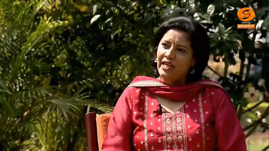 A woman in a red salwar kameez speaks, her expression animated. Lush green foliage forms a backdrop, with a DD Chandana logo visible in the corner.