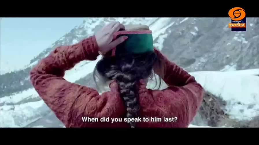 A woman adjusts her traditional cap against a snowy mountain backdrop, the DD Chandana logo visible in the corner. Her braided hair falls down her back as she looks away, a question appearing on screen.