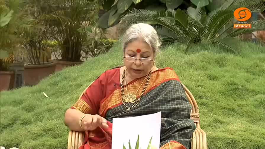 A woman in a red and grey sari speaks while holding a white paper. She wears glasses and a gold necklace, with a red bindi on her forehead.