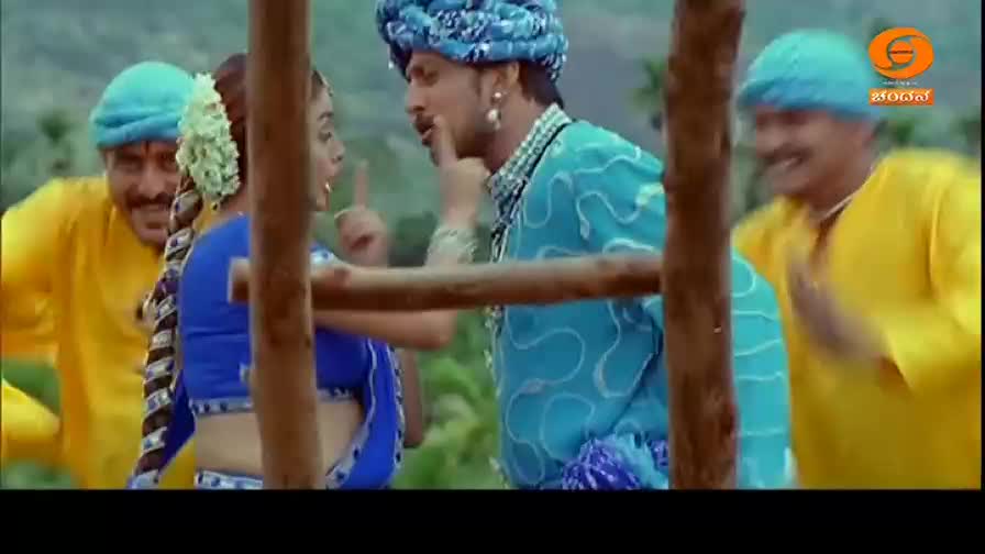 A man in a bright blue turban gestures emphatically at a woman in a blue sari. Another man in a yellow tunic laughs heartily behind them.