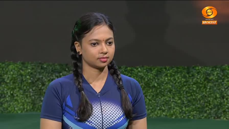 A young woman with dark hair in two braids stands attentively, wearing a blue athletic shirt. A logo for DD Chandana, a television channel from India, is visible in the upper right corner.