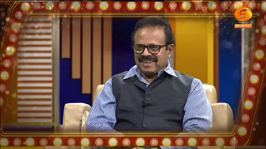 A man with glasses and a mustache sits in a studio chair, smiling. He wears a blue striped shirt and a dark vest, his hands resting on his lap. The backdrop behind him features vertical wooden panels and a bright yellow curtain, typical of a DD Chandana program.