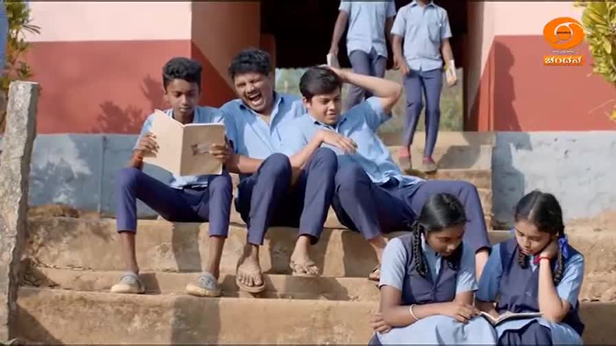 Four students in blue uniforms are gathered on a set of concrete steps. One boy reads from a book while another leans back, laughing heartily. Two girls sit lower down, focused on their own reading.