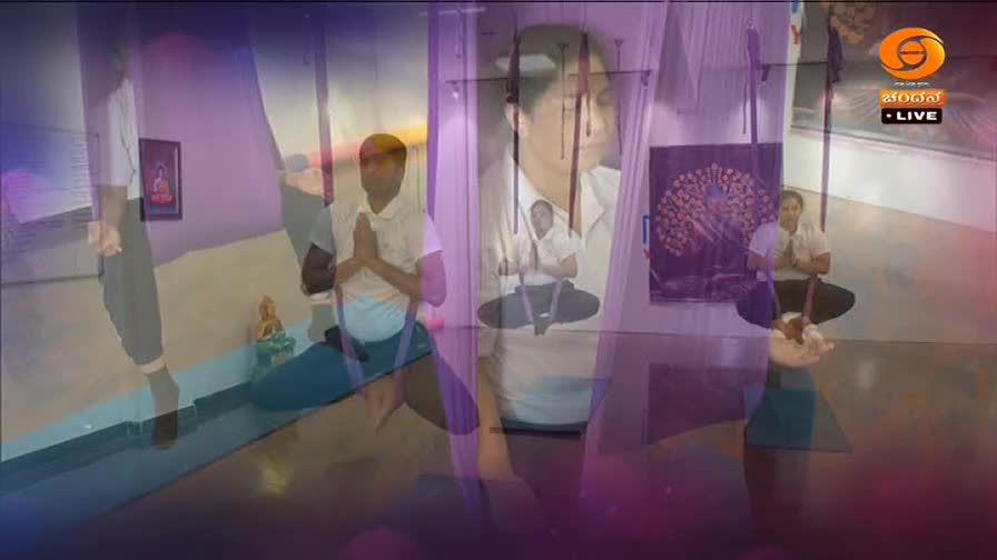 A man in a white shirt and blue pants kneels on a yoga mat, hands pressed together in prayer. Across from him, another person is suspended in a fabric hammock, also in a meditative pose. The DD Chandana logo is visible in the corner.