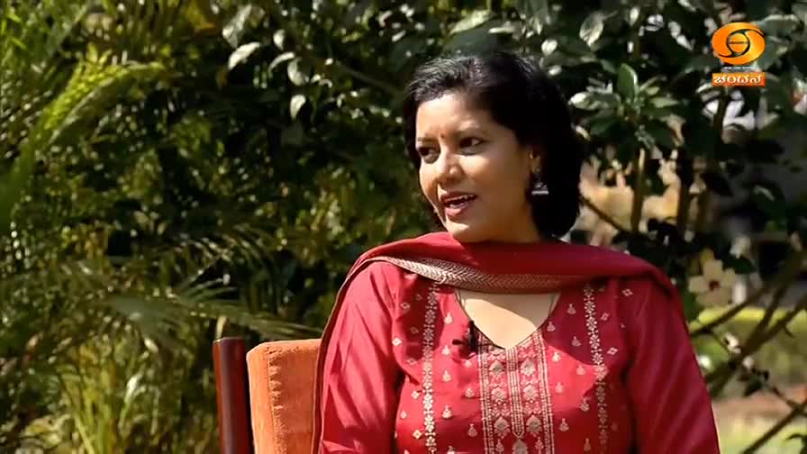 A woman in a red kurta speaks animatedly, framed by lush green foliage. The DD Chandana logo is visible in the upper right corner.