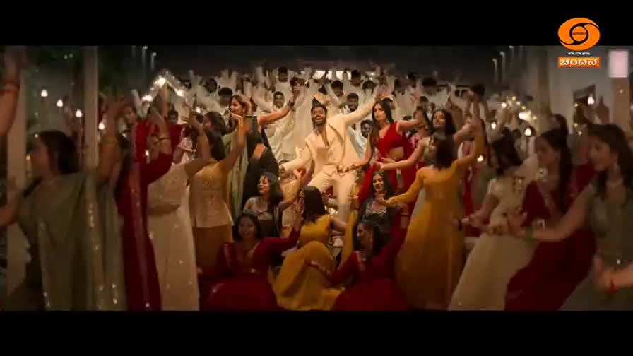 A man in a cream suit is dancing in the center of a large group of women, all dressed in vibrant Indian attire. The women are clapping and moving their hands in unison, their joy evident. A man in a cream suit is dancing in the center of a large group of women, all dressed in vibrant Indian attire. The women are clapping and moving their hands in unison, their joy evident.
