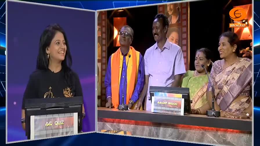 A woman in a black t-shirt smiles brightly, standing behind a podium labeled "Quiz." To her right, a man in a purple shirt and another in a vibrant orange and blue shawl stand with a group of women, all looking towards the stage. The DD Chandana logo is visible in the background, indicating a broadcast from India.